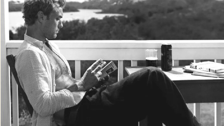 A man reading on a balcony above a wide green landscape, a drink on the table beside him