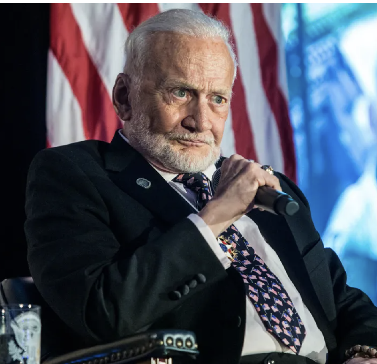 A photo of astronaut Buzz Aldrin in a suit and tie holding a microphone in front of an American flag, used in YouTube videos that claim he said something he never said