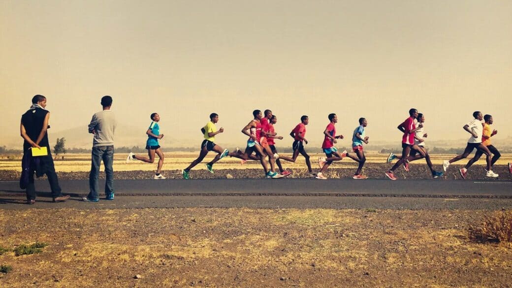 A pack of runners strides along a country road while two men stand at the roadside watching