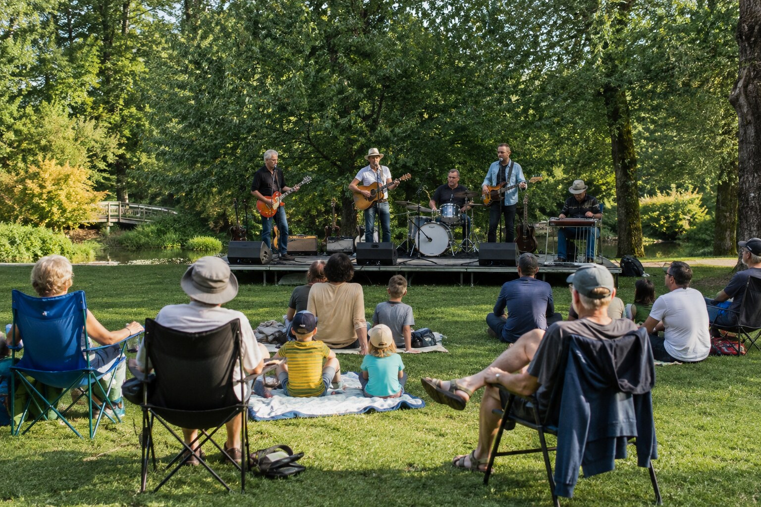 Five-piece band performing in a leafy park for families seated on blankets and chairs