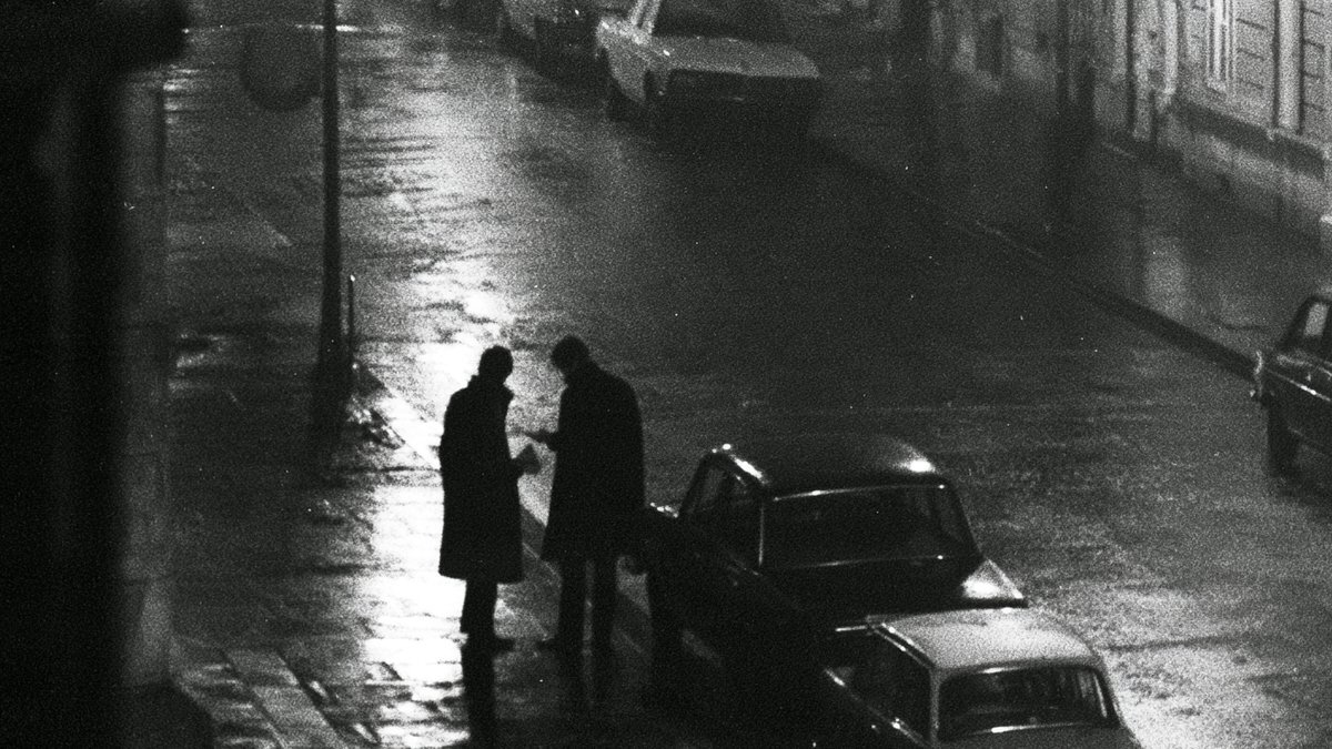 Two figures in long coats exchanging something on a rain-soaked street at night