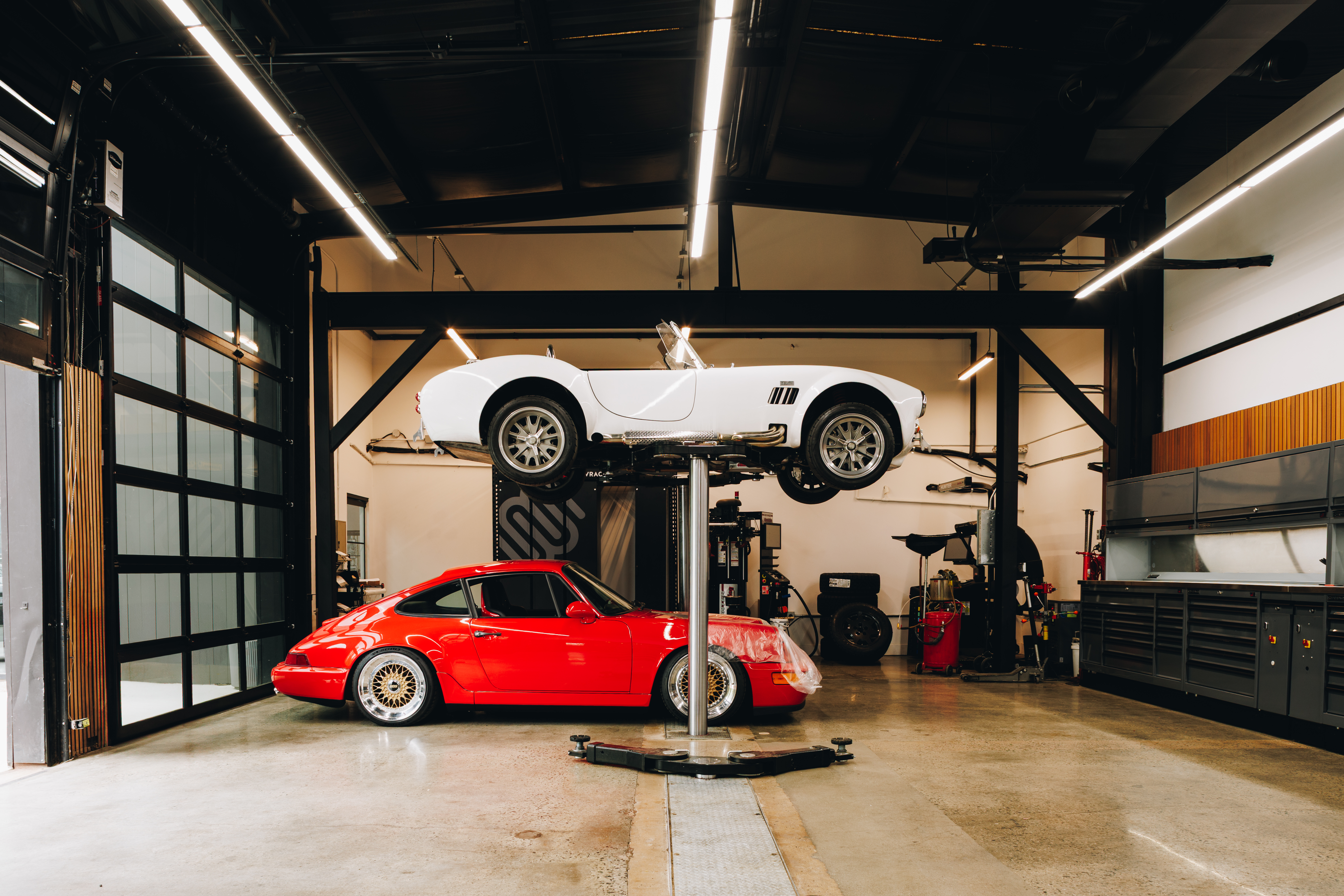 Red Porsche 911 964 below a white Cobra roadster on a lift, Charlotte performance shop