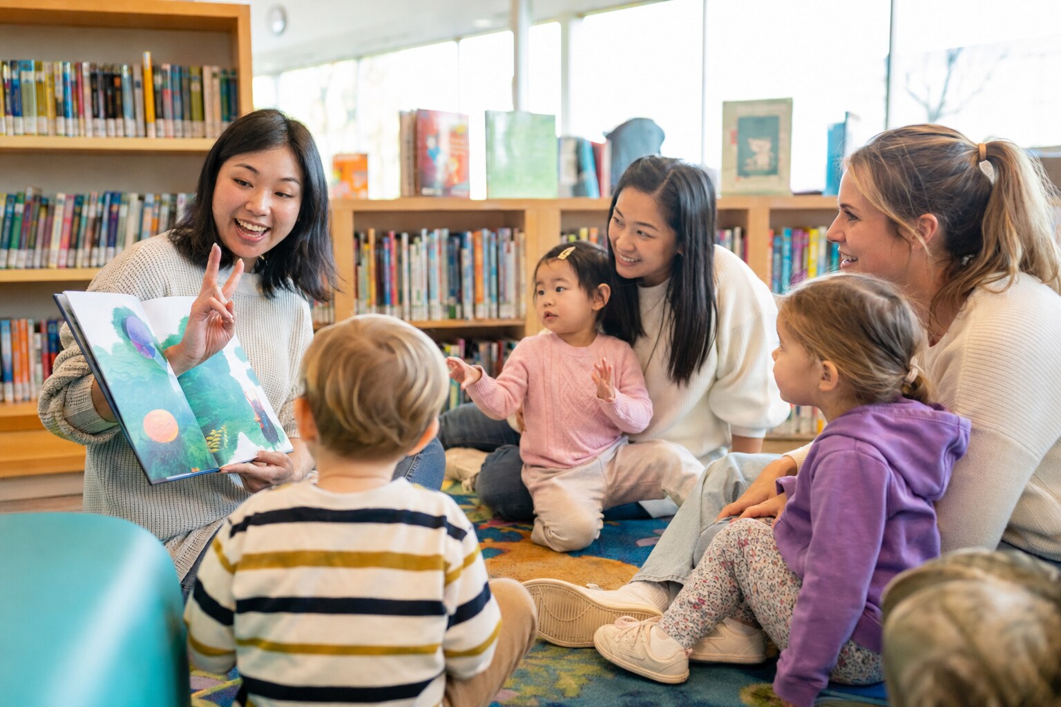 Librarian reading a colourful picture book to toddlers and caregivers seated together on a rug