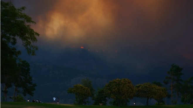 Smoke rising from a wildfire on a ridge, with two figures on parkland in the foreground