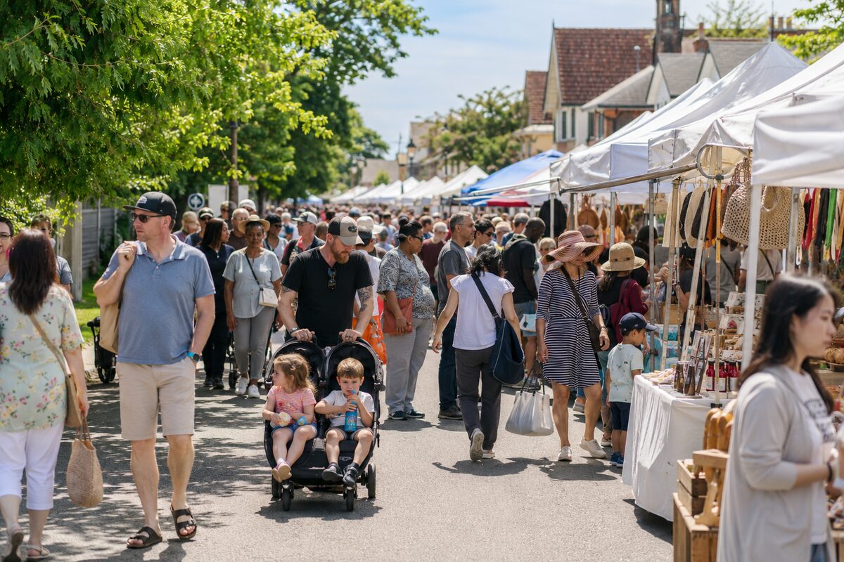 Crowds walking between artisan and food stalls at a sunny open-air village market
