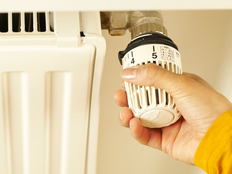 A hand adjusts the numbered thermostatic valve on the side of a white panel radiator.