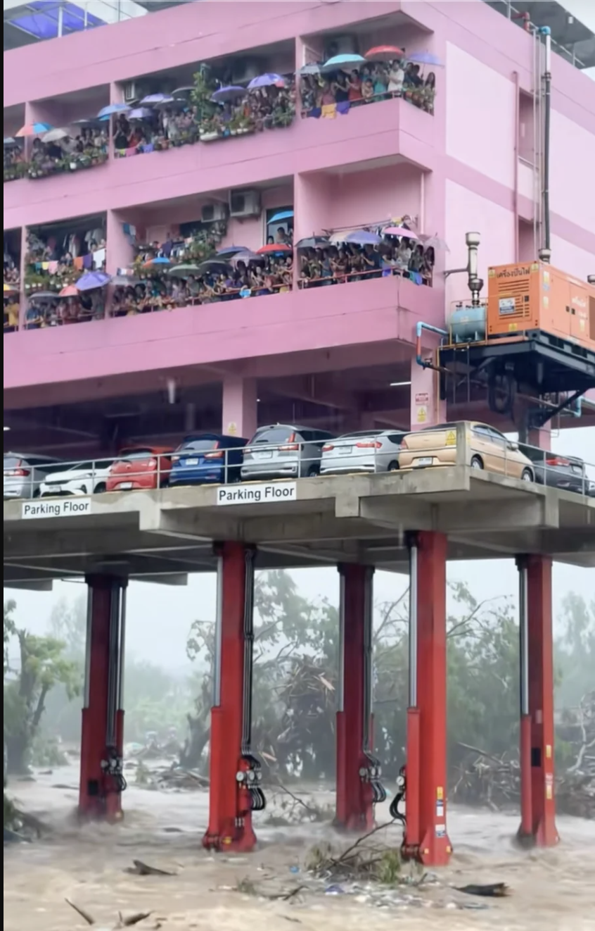 A house lifted above muddy floodwater on bright red hydraulic metal legs while debris rushes underneath, a still from an AI-generated video