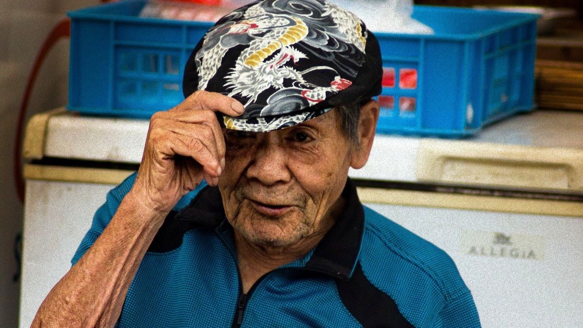 An elderly Japanese man in a patterned cap tipping it towards the camera