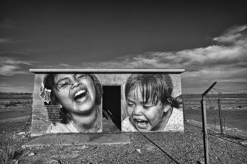 Black-and-white photograph of a small concrete building in a flat desert with a mural of two laughing children, one with a flower in her hair, and an open doorway between them.