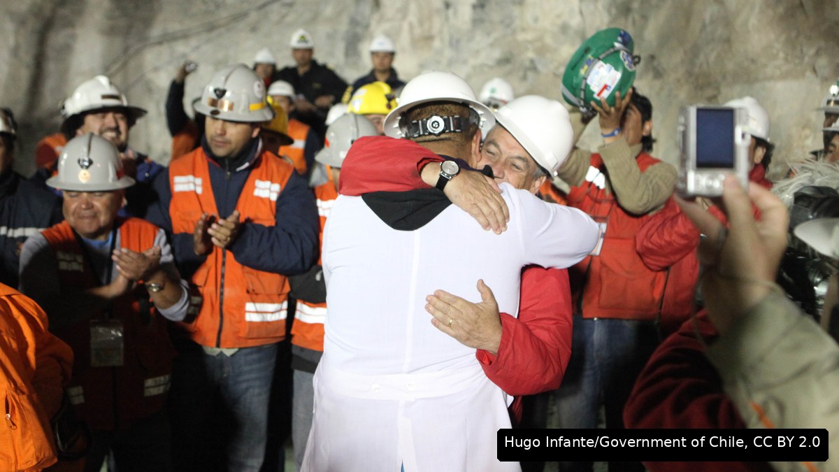 A rescued Chilean miner hugging a man in a hard hat as workers in hard hats and hi-vis cheer around them