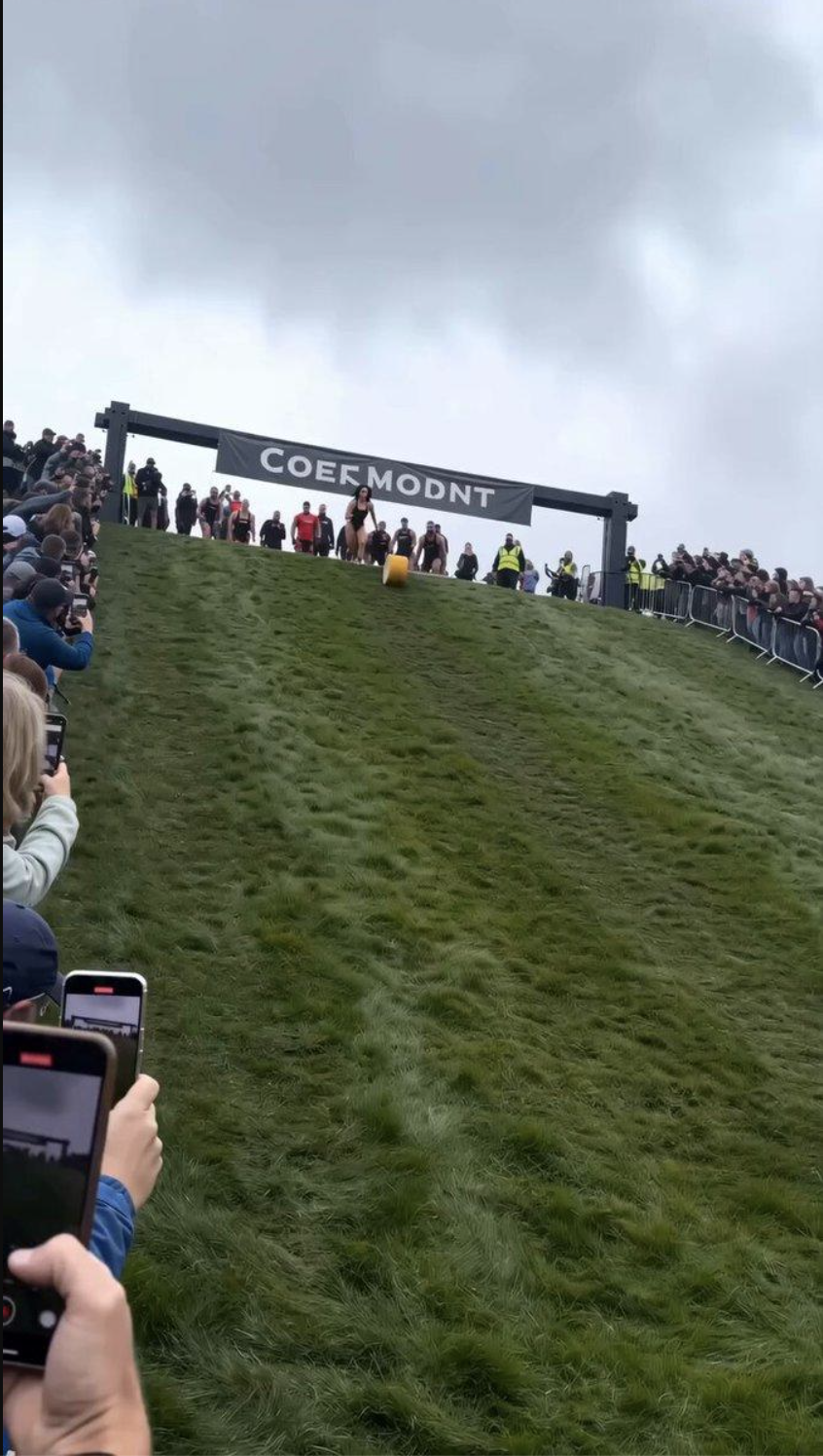 A woman in a swimsuit sprinting downhill after a rolling wheel of cheese, framed as footage of a real competitive race that never actually happened