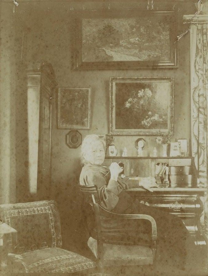 An older woman seated at a desk in a living room hung with framed paintings, in a sepia photograph