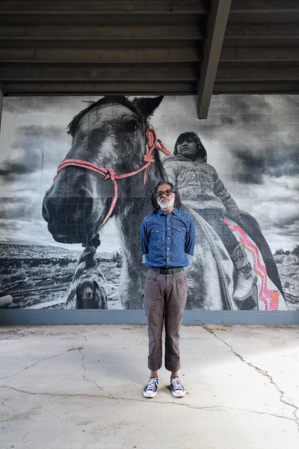Photograph of a bearded man in a denim shirt and sneakers standing in front of a large black-and-white mural of a horse with a pink halter and a rider