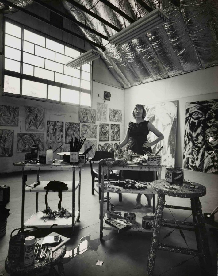 Photograph of a woman standing with hands on her hips in a large studio with tall windows, paint carts, brushes and big drawings on the walls