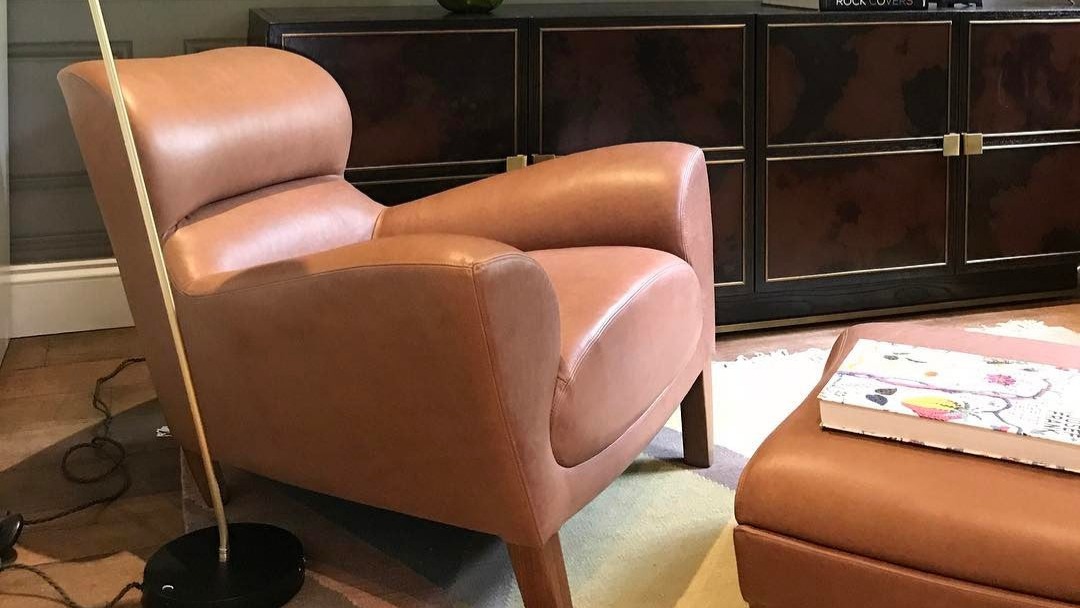 A tan leather reading chair and footstool beside a floor lamp and patterned wallpaper.