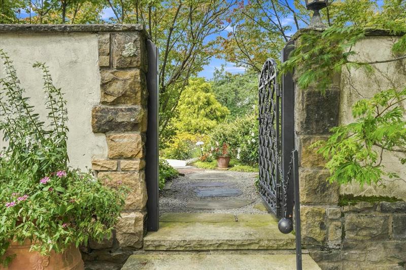 A wrought-iron gate standing open in a stone and stucco wall at 406 Hillside Drive, a flagstone path leading through gravel toward gardens, a potted geranium and Japanese maples