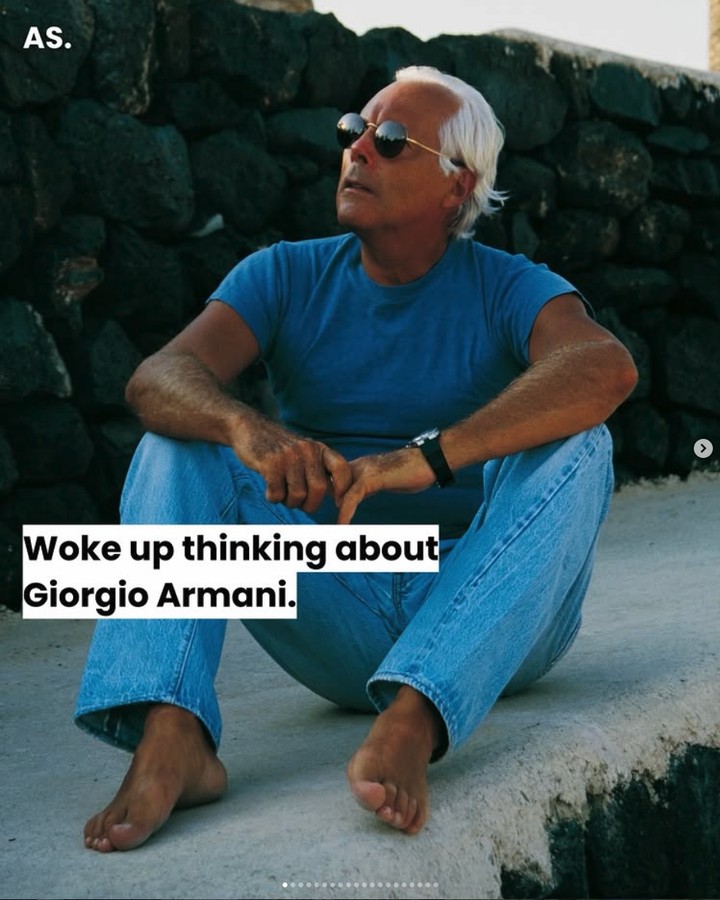 A man in a blue t-shirt and jeans sitting barefoot against a stone wall in the sun