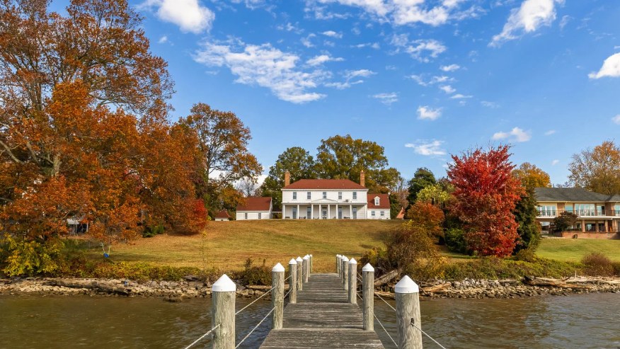 A white colonial house on a lawn above a wooden dock, framed by autumn trees