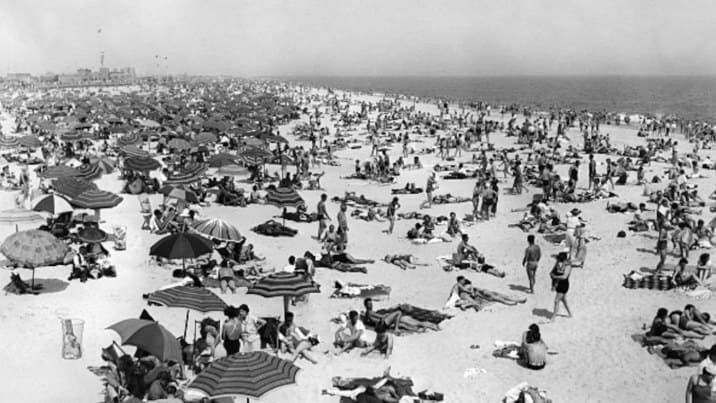 A packed New York beach in the 1930s, sunbathers and striped umbrellas stretching to the horizon