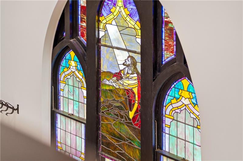A tall arched stained-glass window inside 201 Frederick Avenue, its center panel showing a robed figure kneeling on a rocky hillside in yellow and red glass, flanked by two narrower green and blue panels in dark wood frames against a cream wall