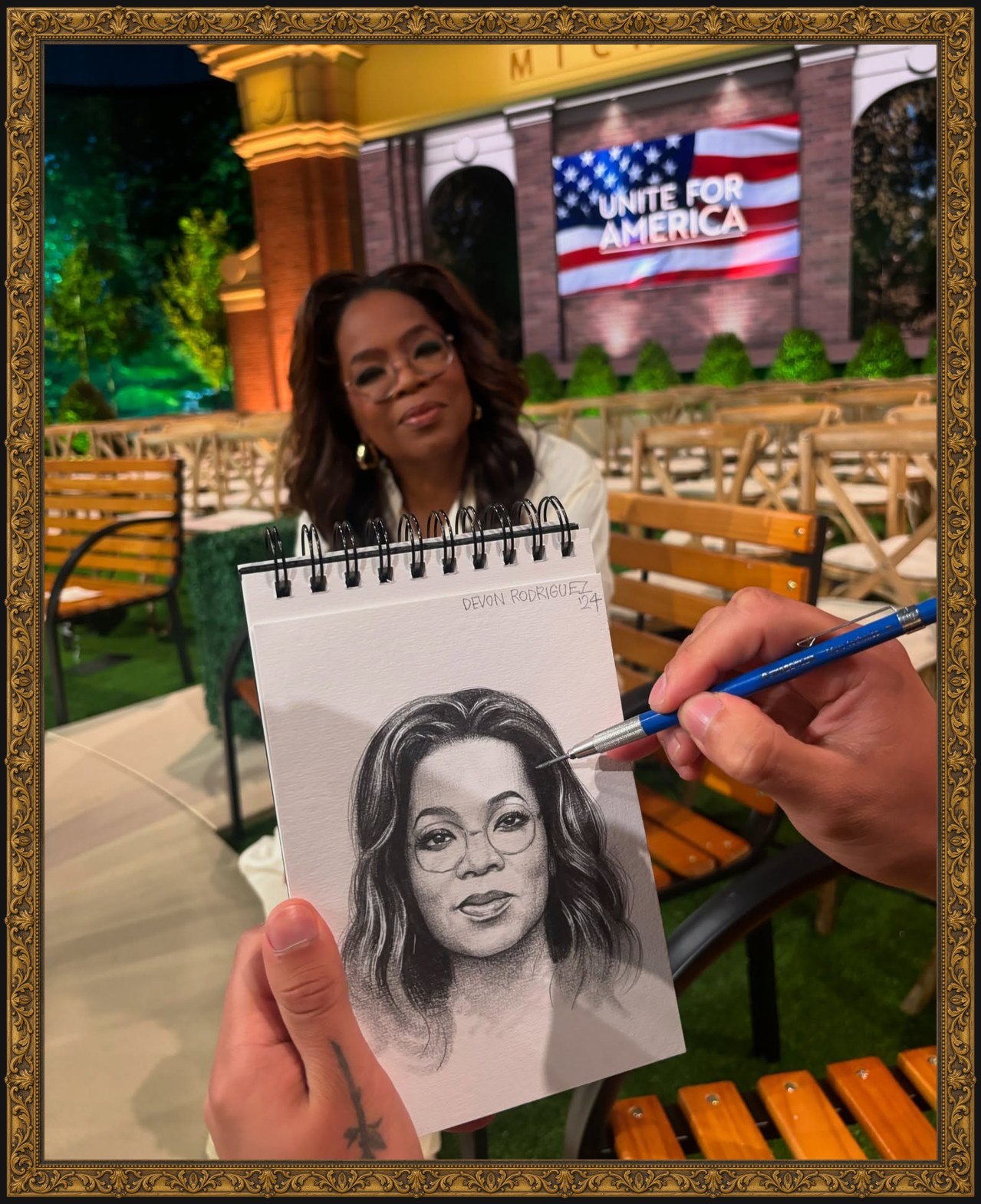 Photograph of a hand holding a small spiral sketchbook with a pencil portrait of a woman with glasses, while the woman sits blurred behind it among wooden chairs