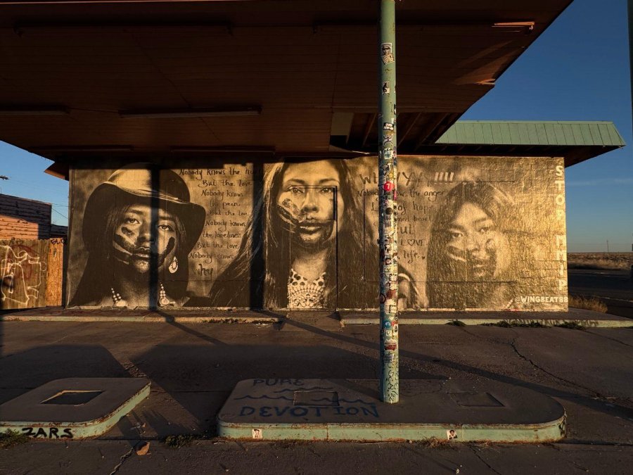 Photograph of a mural on a gas station wall showing three Indigenous women with painted hands across their faces, with handwritten text, in sepia tones under a blue sky.