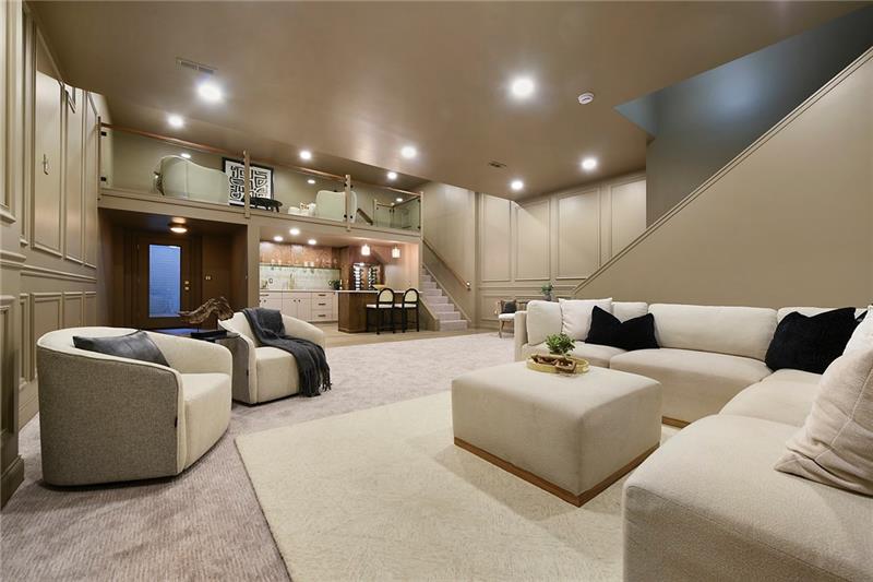The finished lower level at 40 Lemoyne: a bar with stools beneath a glass-railed loft, wainscoted walls, and a sectional around an upholstered ottoman