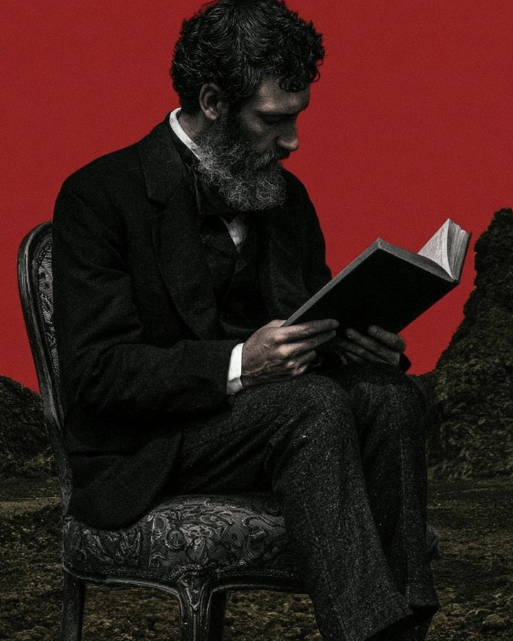 A bearded man in a dark suit reading a book on a chair against a deep red sky