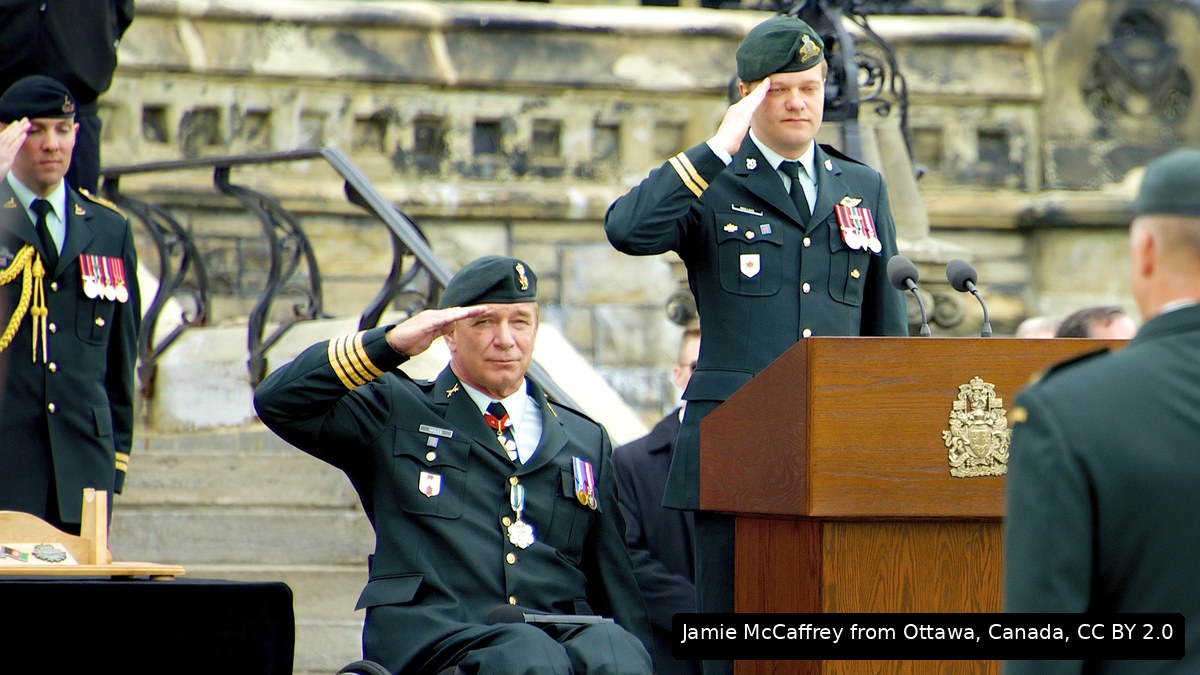 A man in a wheelchair in dress uniform saluting at a ceremony, beside an officer at a lectern