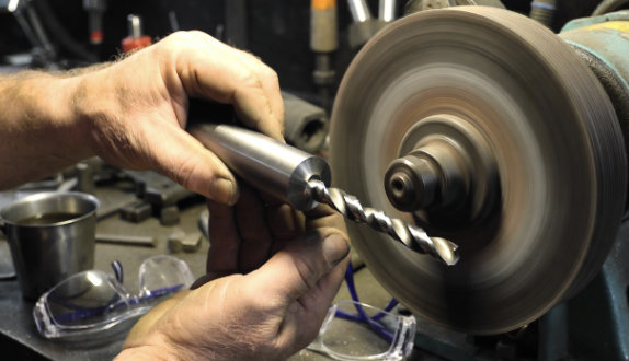 Hands holding a drill bit near a bench grinder with safety glasses nearby