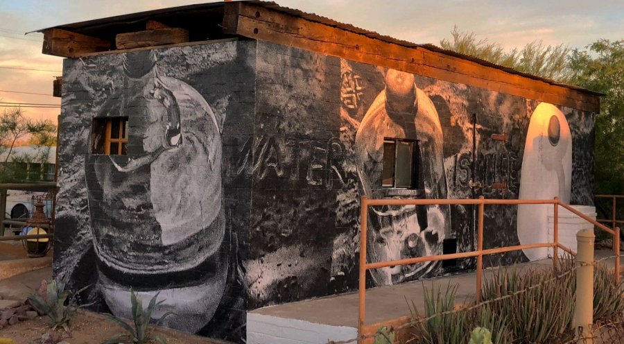 Photograph of a small brick building covered in a black-and-white mural of large water vessels and the words water and life, with an orange railing and cactus in front.