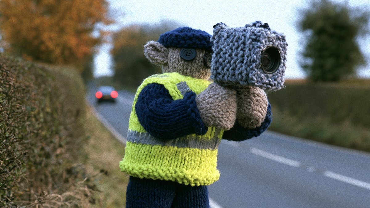 Knitted traffic officer in a high-vis vest holding a knitted speed camera beside a country road