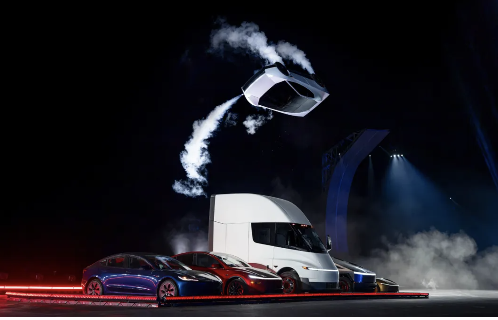A prototype Tesla Roadster flipping in mid-air above a row of parked Tesla vehicles at a live outdoor event in Texas