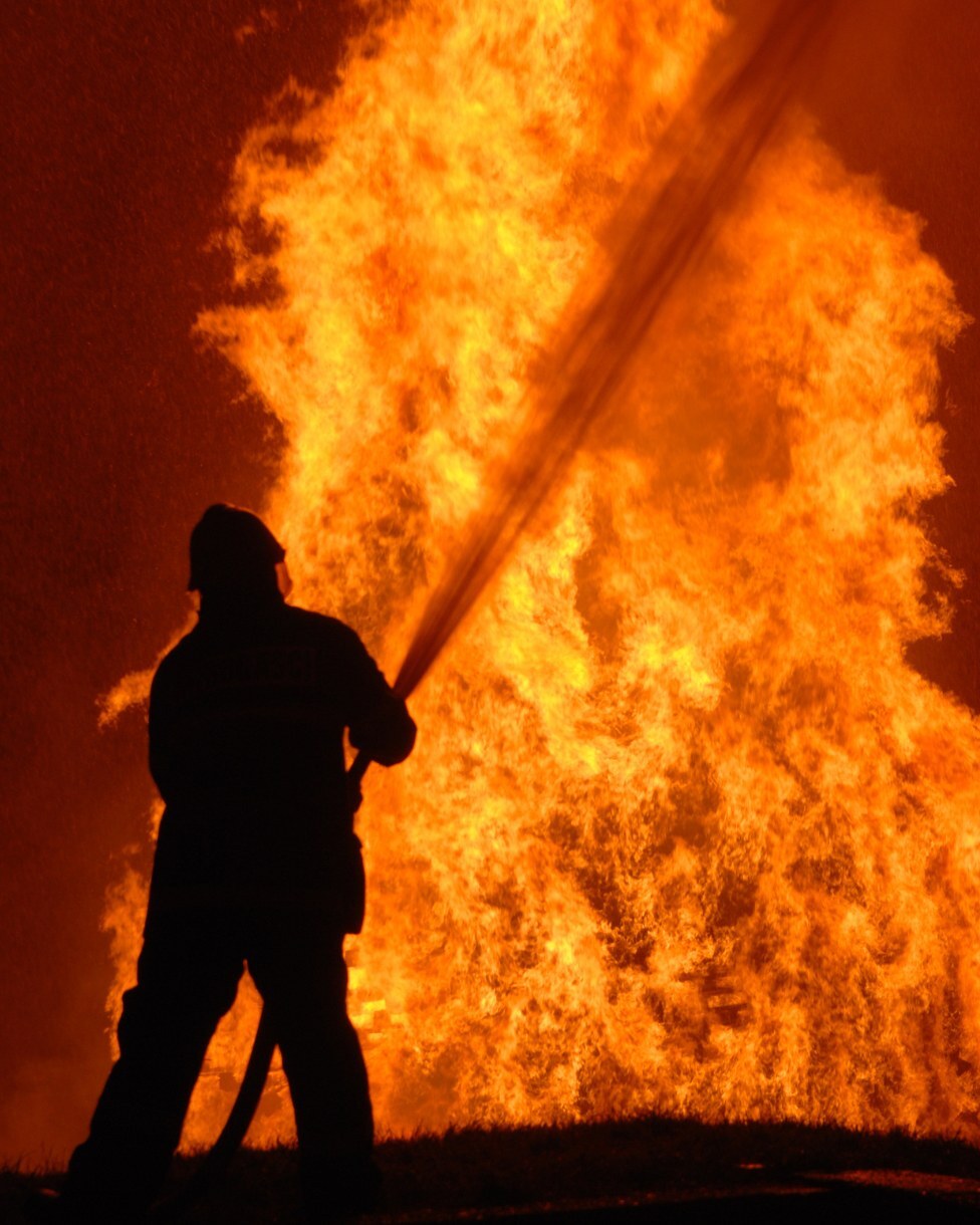 Silhouette of a firefighter spraying a hose at a towering wall of flames