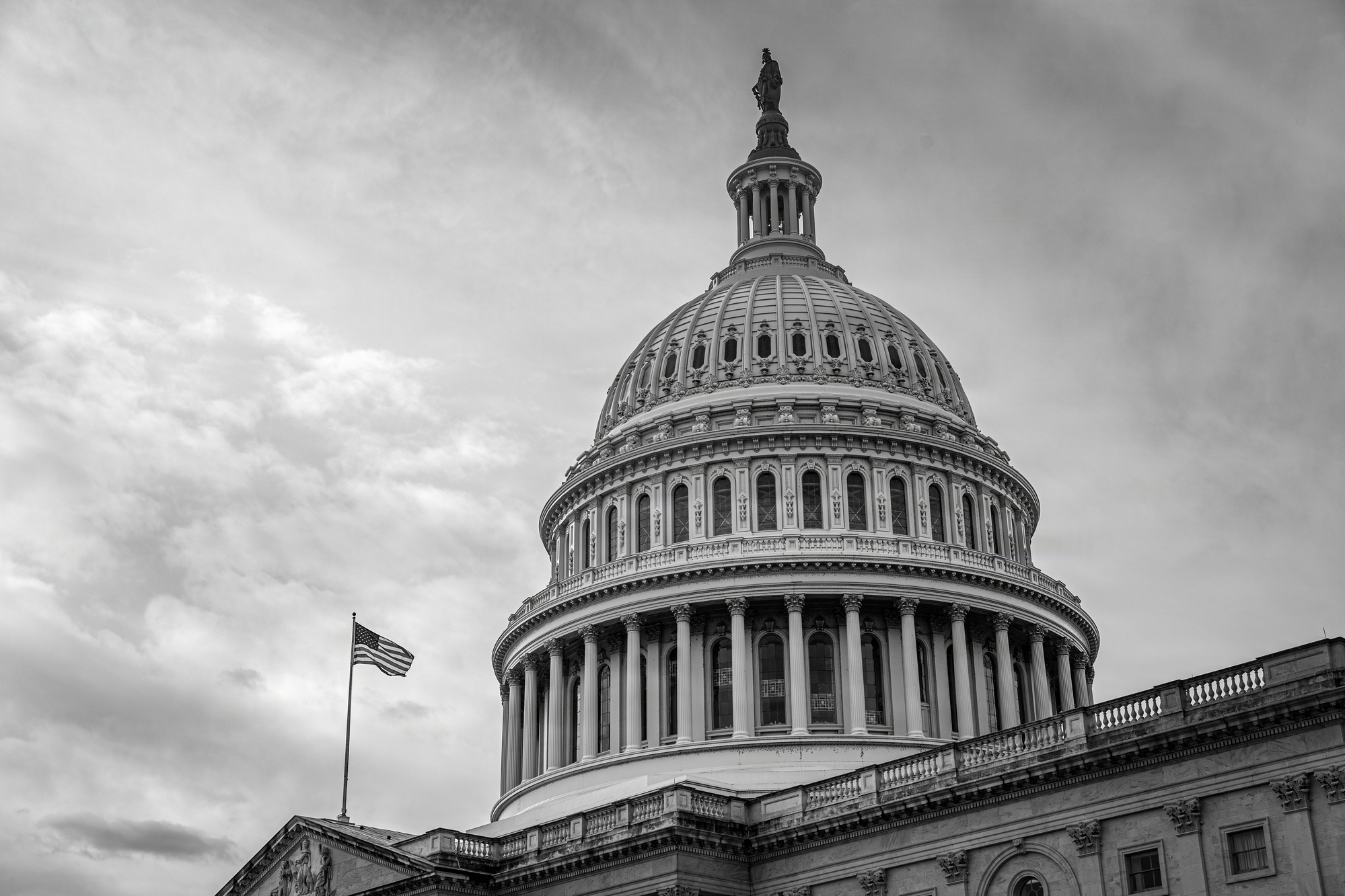 The dome of the United States Capitol under a heavy overcast sky, an American flag flying from the roof below it