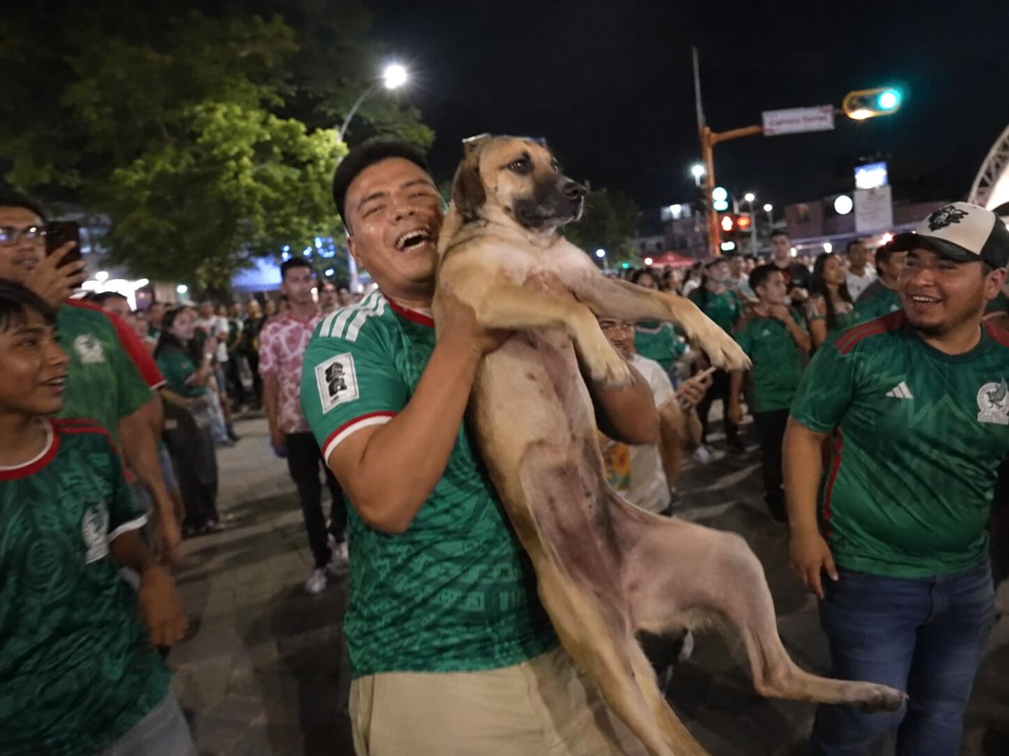 Editorial recreation of a tan dog being held by a smiling Mexico soccer fan during a nighttime street celebration.