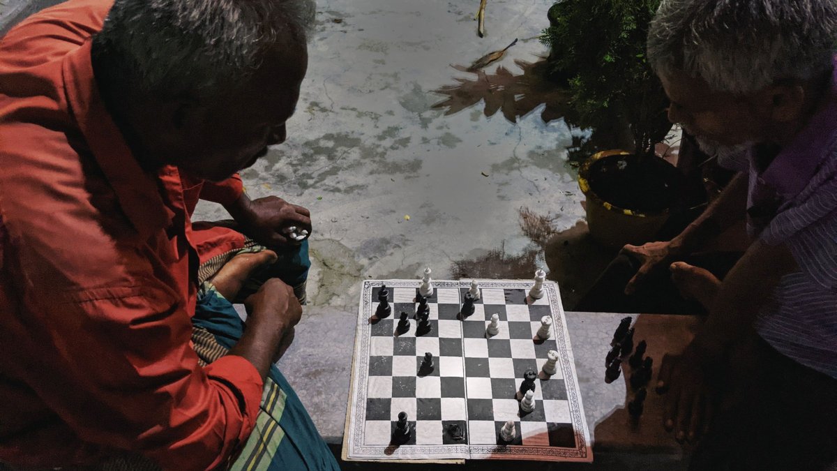 Two older men playing chess on a paper board laid on an outdoor ledge