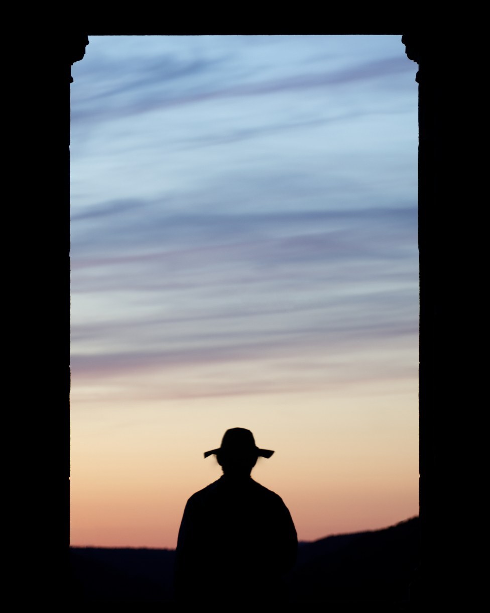 Silhouette of a man in a wide-brimmed hat framed by a dark window against a pastel sunset sky
