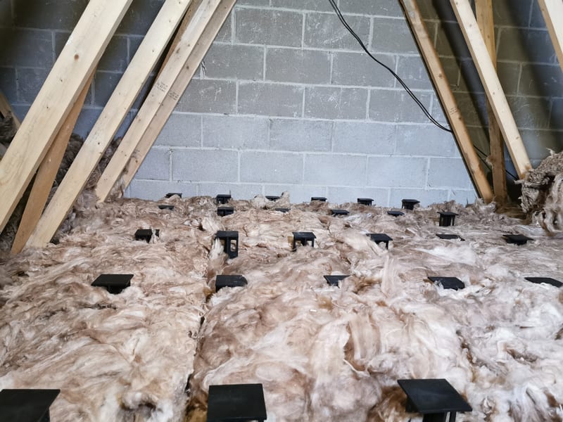 Mineral-wool insulation around black raised-floor supports beneath timber roof trusses and a grey block gable wall.