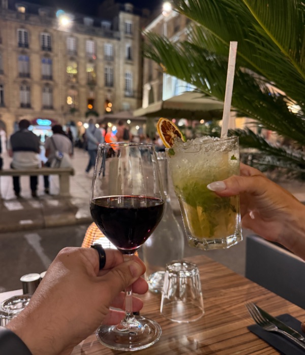 A glass of red wine and a mojito clinking over a sidewalk table at night, with people walking past stone buildings behind them.