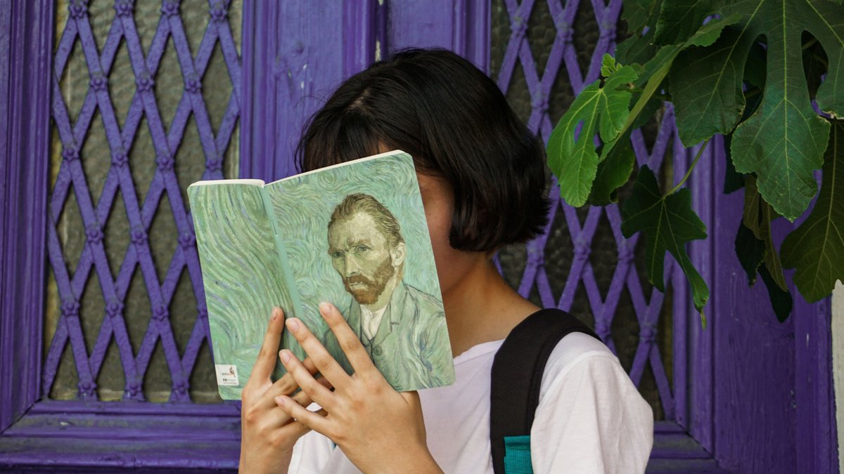 A person holding an open book with a Van Gogh self portrait covering their face, in front of a purple background