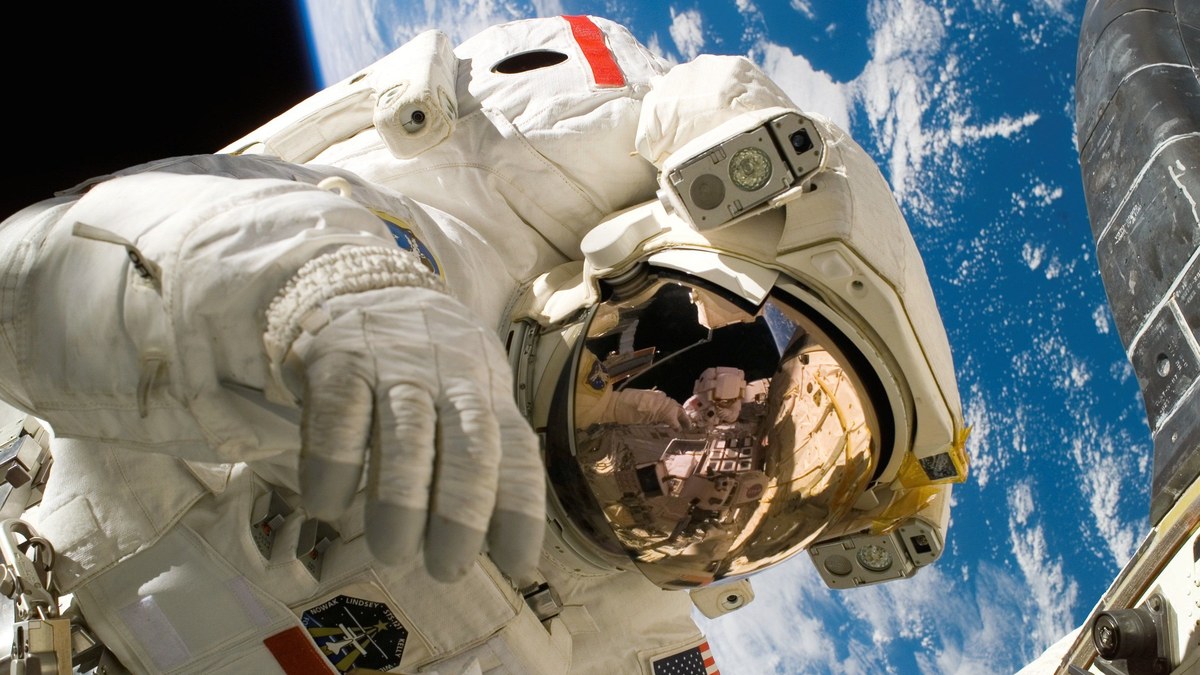 An astronaut on a spacewalk, with the shuttle reflected in the visor and Earth behind