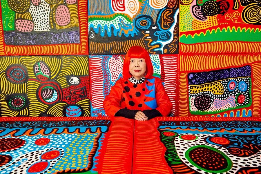 Kusama in a red wig and red coat seated among her brightly patterned paintings
