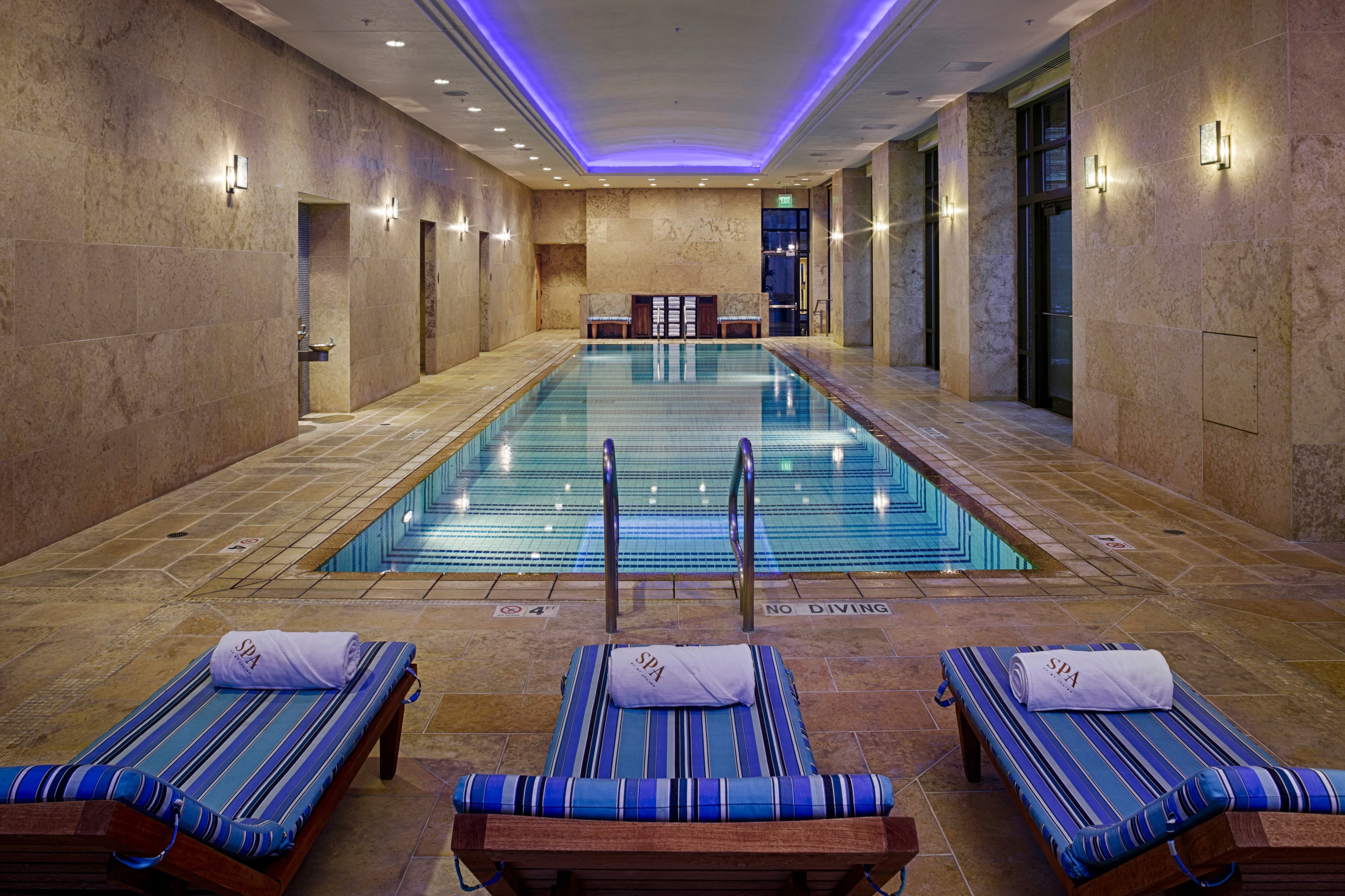 Indoor lap pool lined with loungers beneath violet ceiling lighting