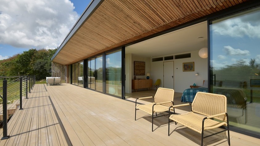 Sliding glass doors opening from a timber-clad home onto a deck with woven chairs.