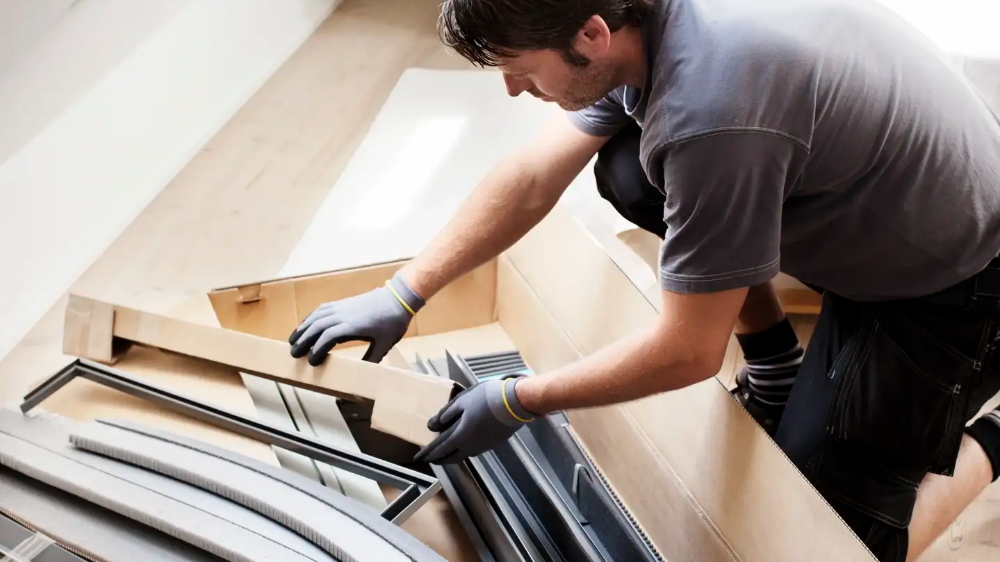 A worker unpacking roof-window components in a bright attic room.