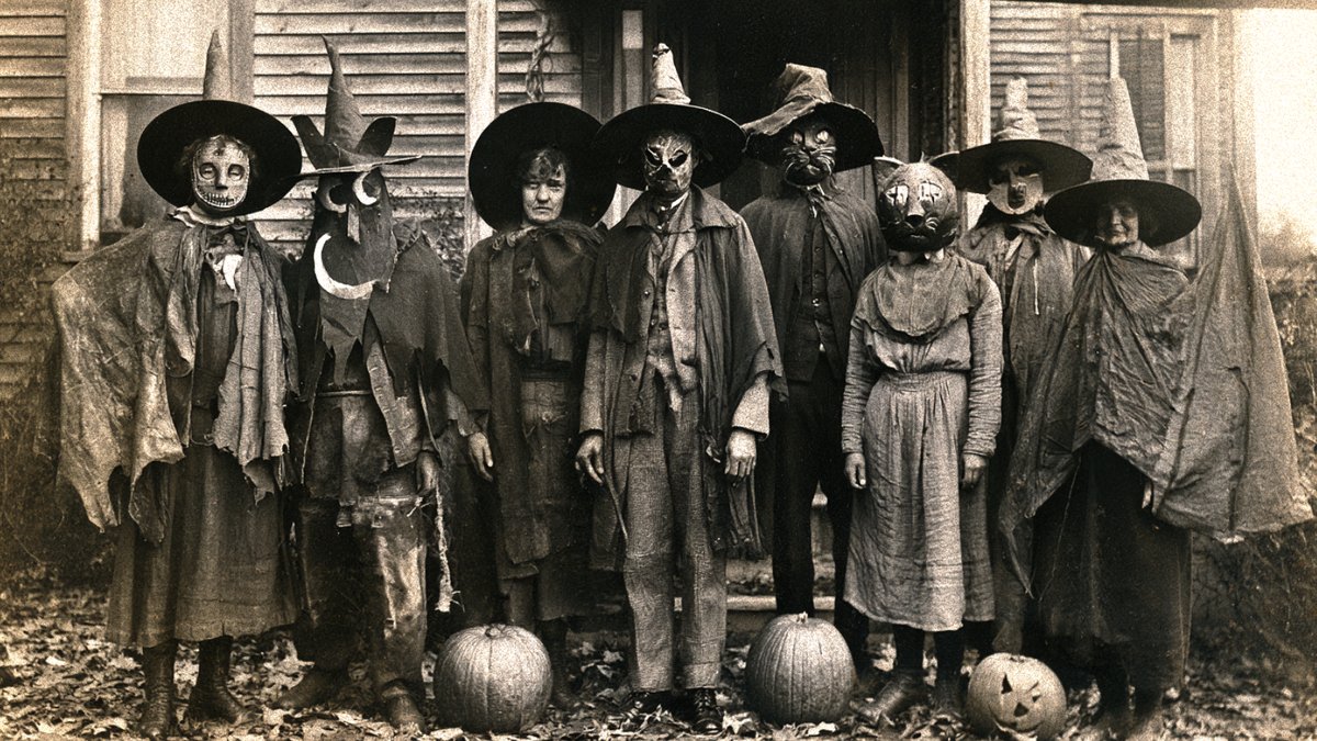 A vintage sepia photo of a group in homemade witch costumes and masks standing behind pumpkins