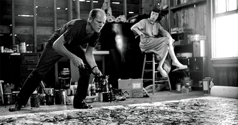 Black and white photograph of a man leaning over a paint-splattered floor canvas holding a paint can, while a woman sits on a ladder stool behind him in a wooden studio
